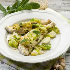 Dwa filety miruny żywej, pieczone z porem i pistacjami, podane na białym talerzu z widelcem i ziołami.