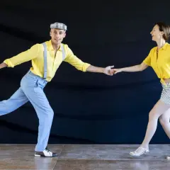 Coppia in abiti vintage che esegue un energico lindy hop ballo, con lui in giallo e bretelle, lei in giallo e gonna a righe.