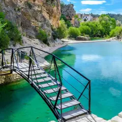 Puente metálico sobre aguas turquesas en uno de los lagos en Valencia, rodeado de rocas y vegetación.