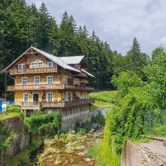 Drewniany pensjonat w górach, otoczony zielenią, z balkonami i strumieniem. Idealne miejsce na rodzinne wakacje, oferujące wiele atrakcji dla dzieci.