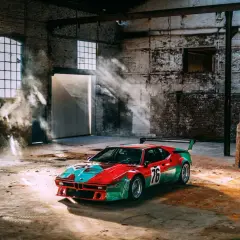 Un m1 coche de carreras con librea roja y verde, número 76, descansa en un almacén polvoriento con rayos de sol y humo.