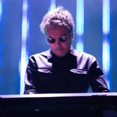 Jean Michel Jarre w Polsce, w okularach przeciwsłonecznych, gra na syntezatorze na tle niebieskich świateł.