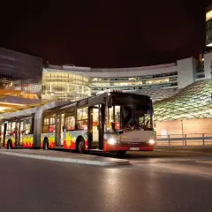 Długi, przegubowy autobus linii 59 w Łodzi nocą, zatrzymuje się przy nowoczesnym centrum handlowym.