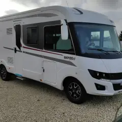 Nowy, biały kamper Rapido gotowy na przygody. W tle widać dom i pomarańczowy miniładowarkę.