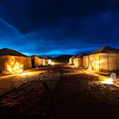 Campement de tentes illuminées dans le désert marocain, sous un ciel nocturne bleu profond. L'ambiance est magique.