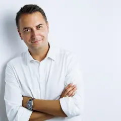 Mężczyzna w białej koszuli, z rękami skrzyżowanymi na piersi, prezentuje pewne siebie pozy do zdjęć męskie. Uśmiecha się lekko do obiektywu.