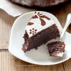 Una fetta di torta caprese ricetta originale napoletana, cosparsa di zucchero a velo e cacao, su un piatto bianco a forma di cuore.