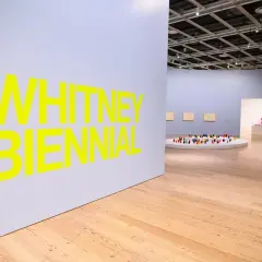 Bright yellow text announces the Whitney Biennial 2026. Inside the gallery, a circular display holds a colorful collection of small figures.