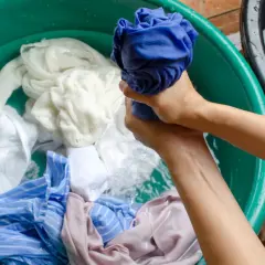 Mains lavant du linge dans une bassine verte. Un vêtement décoloré par le soleil, blanc et bleu, baigne dans l'eau.