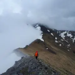 Trekking w Pirenejach. Samotny turysta w pomarańczowym plecaku idzie skalistą granią, otoczony mgłą i ośnieżonymi szczytami.