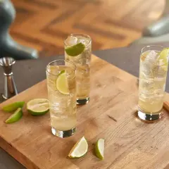 Trzy orzeźwiające whisky ze spritem, udekorowane plasterkami limonki, stoją na drewnianej desce. Obok leżą świeże kawałki limonki.