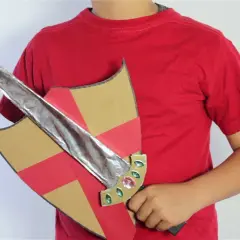 Un niño con camiseta roja sostiene un escudo de cartón con detalles rojos y dorados y una espada de cartón plateada, lista para la aventura.