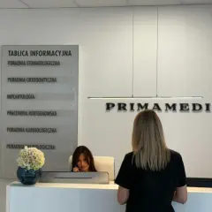 Recepcja kliniki PRIMAMEDICA z tablicą informacyjną i pracownicą.