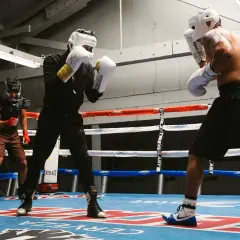 Two boxers in a brawler boxing style spar in a gym, one with white gloves and gold accents, the other shirtless. A third person films them.