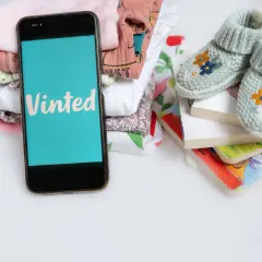 Telefon z aplikacją Vinted obok stosu ubranek dziecięcych i książeczek. Idealne miejsce, by dowiedzieć się, jak kupować na Vinted.