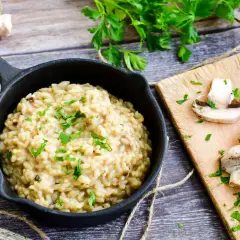 Kremowe risotto z warzywami, posypane natką pietruszki, obok świeże pieczarki na desce.