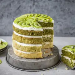 Eine mehrstöckige Matcha Torte mit Kiwi-Scheiben und Sahne. Ein Stück wurde bereits auf einem Teller serviert.