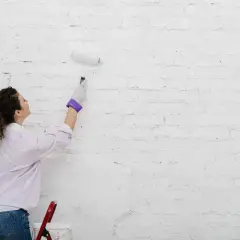 Kobieta w rękawiczce maluje białą ceglaną ścianę wałkiem.