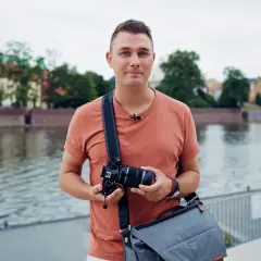 Mężczyzna z aparatem w rękach, na tle rzeki i zabytkowych budynków. Delikatny lens flare dodaje zdjęciu artystycznego charakteru.
