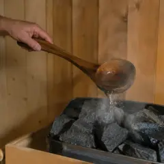 Ein Holzlöffel mit Wasser wird über heiße Steine gegossen, um Dampf zu erzeugen. So kann man sich den Duft von saunahonig selber machen.