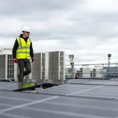 Pracownik w kamizelce odblaskowej przechodzi obok rzędu klimatyzatorów na dachu, gdzie instalowane są panele słoneczne. sofar solar polska.