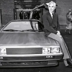 Mężczyzna w garniturze siedzi na masce kultowego **DeLorean**a z otwartymi drzwiami. W tle widać ludzi i budynek.
