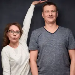 Kobieta z wyrazem zaskoczenia na twarzy, z ręką uniesioną do góry, obok wysokiego mężczyzny. Może to być wizualizacja różnic wzrostu, czasem kojarzonych z zespołem Marfana.