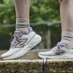Scarpe da tennis New Balance in beige e viola, con calzini beige arricciati, su uno sfondo verde sfocato.