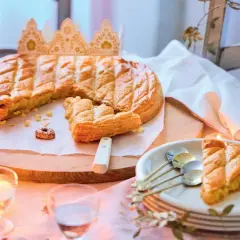 Une galette des rois dorée, prête à être partagée, avec une couronne posée à côté. Une part est déjà servie, invitant à la fête.
