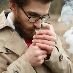 Hombre con barba enciende cigarrillo. El humo forma una calavera, advirtiendo sobre la alergia al humo del tabaco.