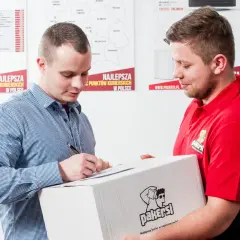 Listonosz w czerwonej koszulce przekazuje paczkę mężczyźnie w kratę, który podpisuje dokument.
