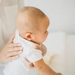 Delikatne dłonie trzymają noworodka w białej koszulce. Dziecko ma lekko pochyloną głowę, a jego twarz jest odwrócona w bok. To typowy obrazek, gdy pojawia się czkawka u noworodka.