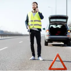Mann mit Warnweste telefoniert neben seinem Auto mit offenem Kofferraum. Ein Warndreieck liegt auf der Straße. Wie viele Warnwesten im Auto sind, ist unklar.