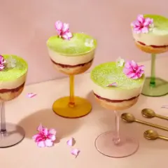 Vackra dessert i glas med lager av kaka, sylt och grön kräm, toppad med blommor. Guldfärgade skedar ligger bredvid.