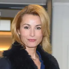 Joanna Brodzik uśmiecha się, ubrana w czarny płaszcz z futrzanym kołnierzem. W tle zielony znak ewakuacyjny.