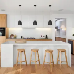 Moderna cocina blanca y madera clara. Isla central con taburetes, electrodomésticos integrados y suelo de madera.