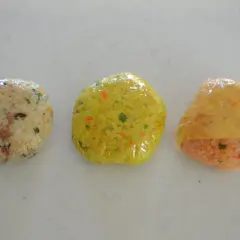 Three plastic-wrapped onigiri sit in a row. The left is white rice with green and brown bits. The middle is yellow rice with orange and green flecks. The right is orange rice with a yellow wrapper. Can you freeze onigiri?