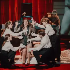 Scena z przedstawienia teatralnego, gdzie aktorzy w białych rękawiczkach otaczają kobietę. To jeden z wielu emocjonujących spektakli, jakie oferują teatry w Polsce.