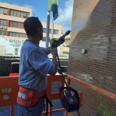 Hombre en plataforma elevadora realiza limpieza de fachadas con agua a presión, dejando la pared de ladrillo brillante y reluciente.