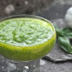 Pesto di basilico bimby in una coppetta, con aglio, pinoli e foglie di basilico sullo sfondo.
