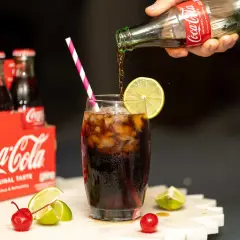 Coca-Cola nalewana do szklanki z lodem i limonką, obok wiśni i cytryn.