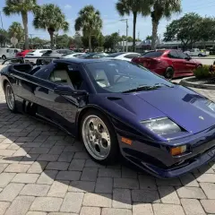 Fioletowy Lamborghini Diablo z jasnym wnętrzem, zaparkowany na brukowanym podjeździe, z palmami w tle.