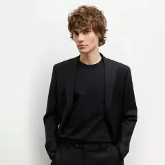 Młody mężczyzna w czarnym garniturze i czarnej koszuli, z rękami w kieszeniach, prezentuje elegancki styl.