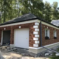 Nowy garaż z cegły, z białym gzymsem i ozdobnymi pilastrami, otoczony zielenią.