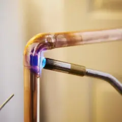 Un chalumeau chauffe un raccord coudé en cuivre pour le souder. La flamme bleue et violette illumine le métal.