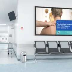 Corridoio di una sala di attesa con sedie, lettini e un monitor che pubblicizza pacchetti di riabilitazione post operatoria.