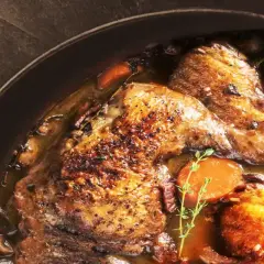 Un plat de coq au vin mijoté, avec des morceaux de poulet dorés, des carottes et des champignons. Le vin rouge utilisé pour la cuisson est essentiel pour ce plat.