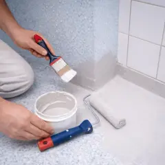 Person målar våtrumsmatta med pensel och roller. En hink med färg står bredvid.