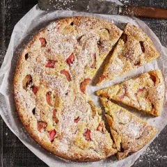 En saftig rabarberkaka, pudrad med florsocker, ligger på bakplåtspapper. Tre bitar är uppskurna.