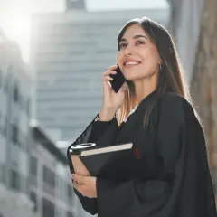 Uśmiechnięta prawniczka w todze rozmawia przez telefon, trzymając książkę i kawę.